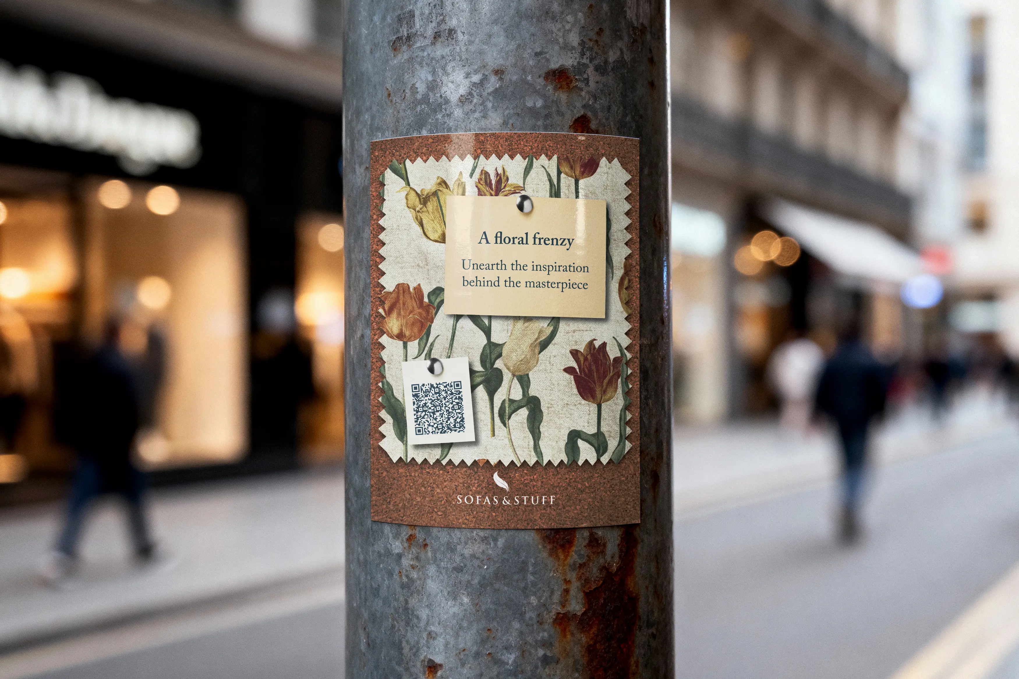 A sticker featuring vintage-style floral art, a QR code, and the text A floral frenzy. Unearth the inspiration behind the masterpiece is attached to a metal pole on a city street with blurred pedestrians and shops in the background.